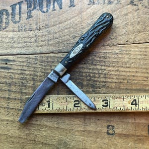 May include: A vintage Case pocket knife with two blades, one partially open. The handle is black with a textured pattern. The blades and metal components show signs of wear. The knife rests on a wooden surface with a ruler.