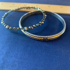 May include: Two silver bangles with gold accents and decorative embellishments. One bangle features small, colorful gemstones. The other has a gold band with a textured design. The bangles are on a blue surface.