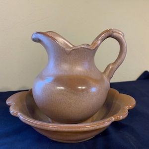 May include: A brown glazed ceramic pitcher and matching bowl. The pitcher has a rounded body and a curved handle. The bowl has a scalloped edge.