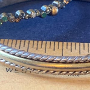 May include: Close-up of a silver and gold bracelet with a beaded accent. The bracelet has a braided design and a gold band. The beaded accent includes clear, green, and gold beads. The word "WEST" is partially visible.