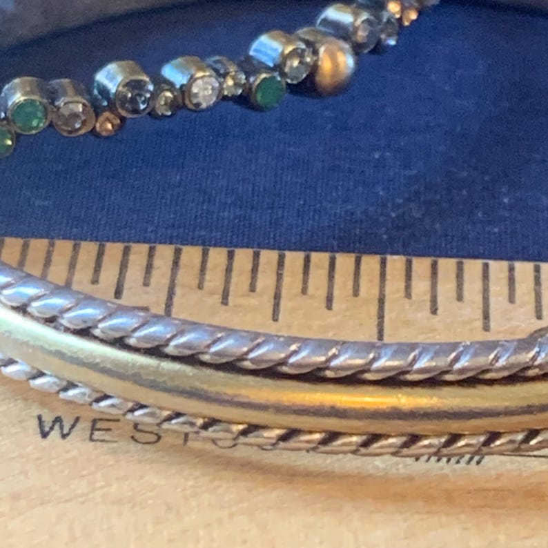 May include: Close-up of a silver and gold bracelet with a beaded accent. The bracelet has a braided design and a gold band. The beaded accent includes clear, green, and gold beads. The word "WEST" is partially visible.