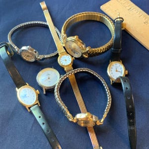 May include: A collection of vintage wristwatches with various band styles, including gold-toned metal, silver-toned metal, and black leather. The watch faces are primarily white with black numerals and hands. A wooden ruler is in the background.