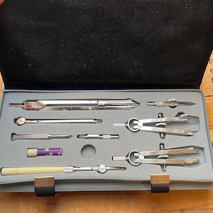 May include: A set of drafting tools in a gray case. The set includes several compasses, dividers, and other drawing instruments. The tools are made of silver metal and are arranged in a foam insert. The case is open, revealing the tools.