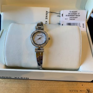 May include: A silver Anne Klein watch with a white face and a bezel encrusted with small, clear crystals. The watch is in a white box with the brand name visible. The band is also silver and has crystal accents.