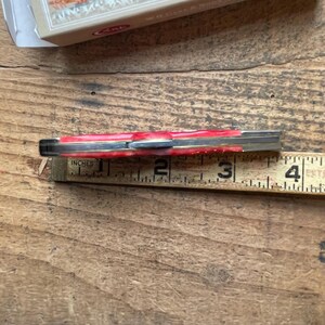 May include: A closed folding pocket knife with a red marbled handle and metallic bolsters, positioned against a wooden ruler showing a length of approximately 3.5 inches, with a partial product box visible at the top of the frame.