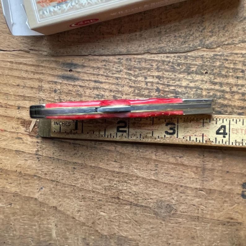 May include: A closed folding pocket knife with a red marbled handle and metallic bolsters, positioned against a wooden ruler showing a length of approximately 3.5 inches, with a partial product box visible at the top of the frame.