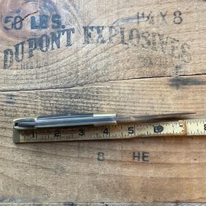 Puede incluir: Una navaja de bolsillo vintage con una hoja parcialmente abierta, junto a una regla de madera. La navaja tiene un mango de lat&oacute;n y una hoja plateada. La regla muestra medidas en cent&iacute;metros.