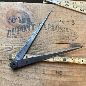 May include: An antique metal compass tool with pointed ends, open on a wooden surface with the text "50 LBS. DUPONT EXPLOSIVES". Two wooden rulers with inch markings are also visible.