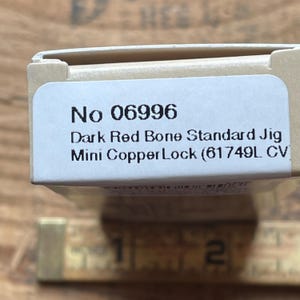 May include: A rectangular product box with a white label featuring the text No 06996 Dark Red Bone Standard Jig Mini CopperLock (61749L CV). A wooden ruler is positioned below the box, showing a measurement of approximately 2 inches.
