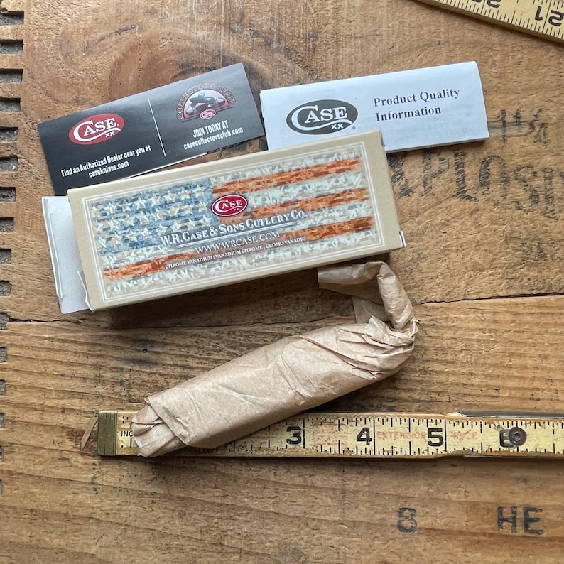 May include: A W.R. Case & Sons Cutlery Co. product box featuring an American flag design, accompanied by a Case XX product quality information card and a collector club card. A brown paper-wrapped item rests above a yellow measuring tape.