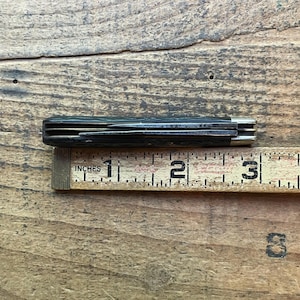 May include: A closed pocket knife with a dark handle and silver accents, resting on a wooden ruler. The ruler measures in inches, with the knife appearing to be approximately 3 inches long.