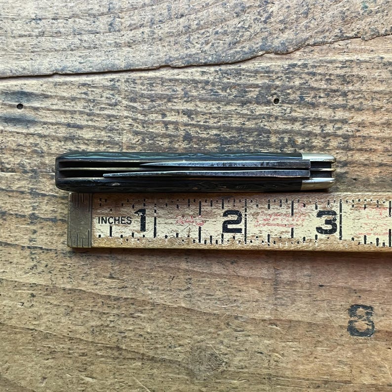 May include: A closed pocket knife with a dark handle and silver accents, resting on a wooden ruler. The ruler measures in inches, with the knife appearing to be approximately 3 inches long.