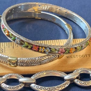 May include: Silver-toned bangles with floral and textured designs. One bangle features colorful enamel flowers with orange, pink, and blue accents. The other bangle has a textured pattern. A silver chain bracelet is also visible.