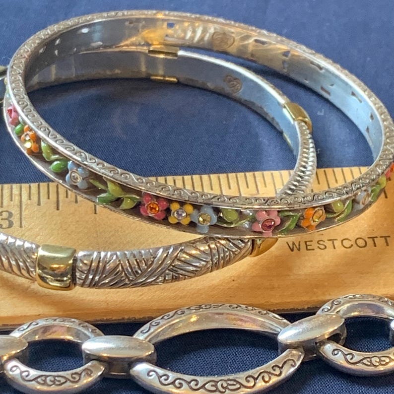 May include: Silver-toned bangles with floral and textured designs. One bangle features colorful enamel flowers with orange, pink, and blue accents. The other bangle has a textured pattern. A silver chain bracelet is also visible.