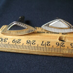 May include: A pair of vintage cufflinks with a gold-tone finish. Each cufflink has a unique design, one with an open, geometric shape and the other with a white, rectangular center. The cufflinks are placed on a wooden ruler.