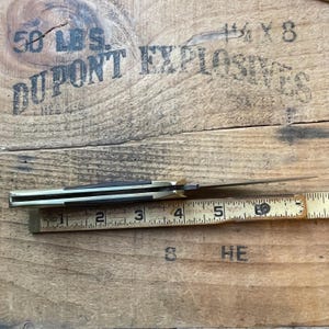 Puede incluir: Una navaja de bolsillo vintage con mango de lat&oacute;n y hoja oscura, mostrada junto a una regla de madera. La regla mide la navaja en aproximadamente 13 cm de longitud. La navaja est&aacute; sobre una superficie de madera.