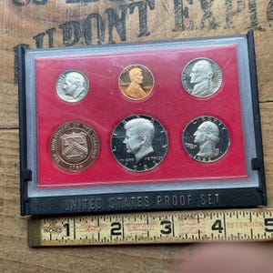 May include: A United States Proof Set in a black plastic case with a red interior. The set includes six different coins, including a penny, nickel, dime, quarter, and half dollar. The case is labeled "UNITED STATES PROOF SET".