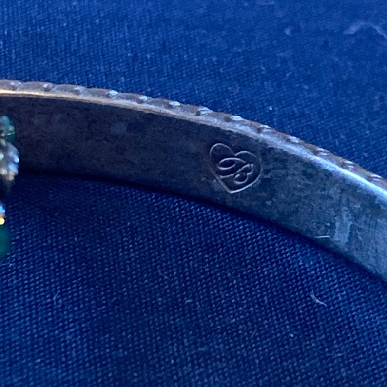 May include: Close-up of a silver-toned bracelet with a decorative emblem. The emblem appears to be a stylized letter "B" within a heart-shaped design. The bracelet has a slightly tarnished appearance, suggesting an antique or vintage piece.