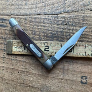 May include: A folding pocket knife with a brown textured handle and silver metal bolsters. The handle features an Old Timer shield. The blade is partially open, revealing the Schrade USA 194T stamp. A wooden ruler shows the knife is 3 inches long.