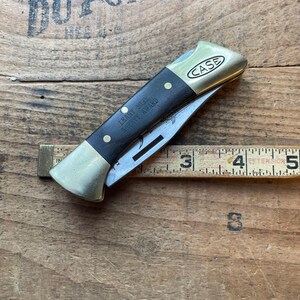 Puede incluir: Una navaja plegable Case vintage con mango negro y detalles en lat&oacute;n. La hoja est&aacute; parcialmente abierta, revelando el filo de acero. El mango est&aacute; inscrito con "LEARY MINE SAFETY AWARD". Una regla de madera est&aacute; junto a la navaja.