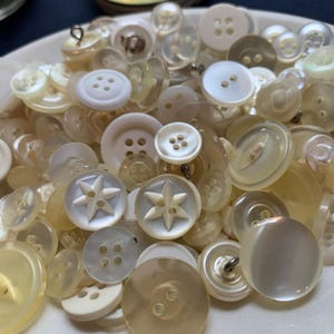 May include: A collection of vintage buttons in various sizes and shapes, in shades of white, cream, and pale yellow. Some buttons feature starburst designs, while others have multiple holes for sewing. The buttons are made of different materials, including what appears to be mother-of-pearl.