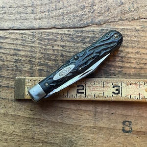 May include: A closed black pocket knife with a textured handle and a partially visible blade. The knife has the word "CASE" on the side and is next to a wooden ruler. The ruler shows measurements in inches.
