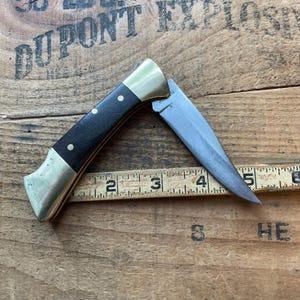 Puede incluir: Un cuchillo plegable parcialmente abierto con mango negro, refuerzos de lat&oacute;n y hoja plateada. El mango tiene tres remaches de lat&oacute;n. El cuchillo est&aacute; colocado sobre una superficie de madera con una regla.