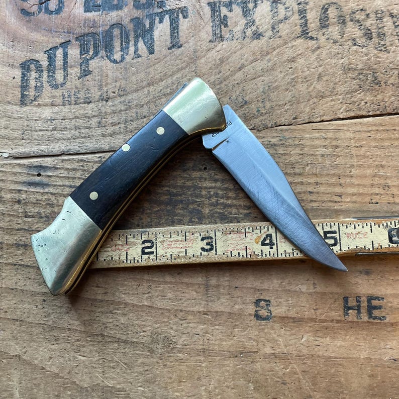 Puede incluir: Un cuchillo plegable parcialmente abierto con mango negro, refuerzos de lat&oacute;n y hoja plateada. El mango tiene tres remaches de lat&oacute;n. El cuchillo est&aacute; colocado sobre una superficie de madera con una regla.