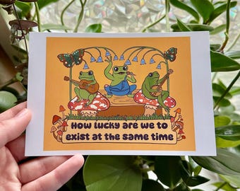 May include: A yellow background with three green frogs playing music on mushrooms. The frogs are wearing overalls, a hat, and a bandana. The text reads "How lucky are we to exist at the same time".