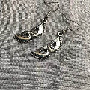 Op de afbeelding: Een paar zilverkleurige oorbellen in de vorm van een masker. Elke oorbel heeft een maskerontwerp met oogopeningen en een decoratieve rand. De oorbellen hebben een haaksluiting voor doorboorde oren en worden gepresenteerd op een grijze stoffen ondergrond.