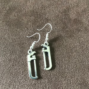May include: A pair of silver-colored earrings featuring a miniature saw design. The earrings have a hook closure and are displayed against a brown textured background. The saw blade is visible in the design.