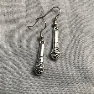 Op de afbeelding: Een paar zilverkleurige microfoon oorbellen. Elke oorbel heeft een gedetailleerd microfoonontwerp met een getextureerde, afgeronde bodem en een cilindrische bovenkant. Het nummer "8" is zichtbaar op de behuizing van de microfoon. De oorbellen hangen aan een eenvoudige haak.