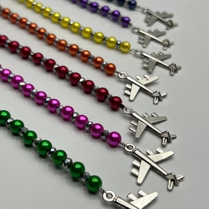 以下が含まれることがあります： 虹色のビーズブレスレットのコレクション。それぞれに銀色の飛行機のチャームが付いています。ビーズは丸く光沢があり、銀色のスペーサーが付いています。ブレスレットは白い背景に対して斜めに配置されています。