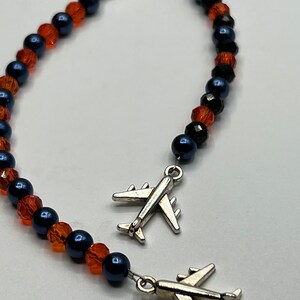 May include: A beaded bracelet with alternating orange and navy blue beads. Two silver airplane charms hang from the bracelet. The bracelet is fastened with a silver clasp. The beads are faceted and round.