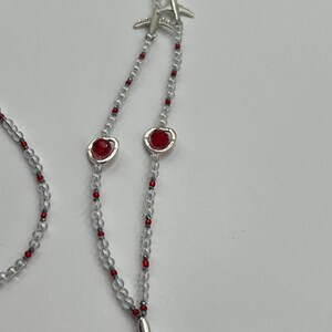 May include: A necklace featuring clear beads interspersed with small red beads. The necklace has silver airplane charms and red heart-shaped charms. The necklace is displayed against a white background.
