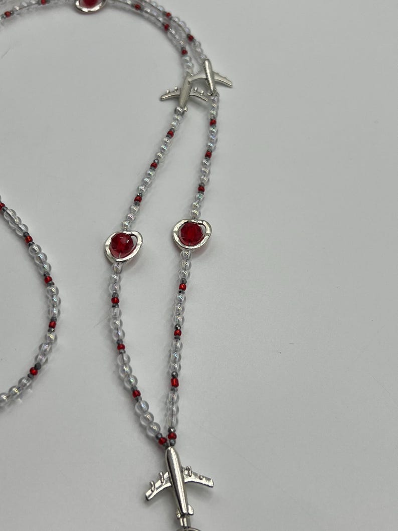 May include: A necklace featuring clear beads interspersed with small red beads. The necklace has silver airplane charms and red heart-shaped charms. The necklace is displayed against a white background.