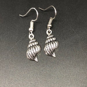 May include: A pair of silver-colored seashell earrings. Each earring features a detailed, textured seashell charm suspended from a simple fish hook ear wire. The earrings are displayed against a dark background.