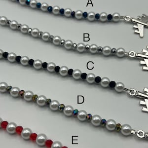 May include: Close-up of five beaded necklaces with airplane charms. Each necklace features white faux pearls and accent beads in various colors: dark blue, silver, black, iridescent, and red. The charms are silver-toned.