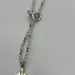 May include: A necklace featuring clear beads with purple accents and a silver airplane charm. Two heart-shaped pendants with purple centers are also present. The necklace is designed with a delicate and feminine aesthetic.