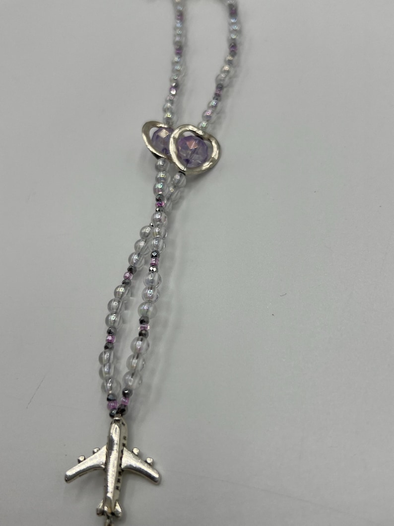 May include: A necklace featuring clear beads with purple accents and a silver airplane charm. Two heart-shaped pendants with purple centers are also present. The necklace is designed with a delicate and feminine aesthetic.