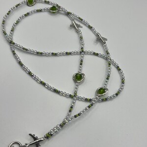 May include: A beaded lanyard with clear, silver, and green beads. The lanyard features silver airplane charms and green heart-shaped beads. A silver clasp is attached at the end.