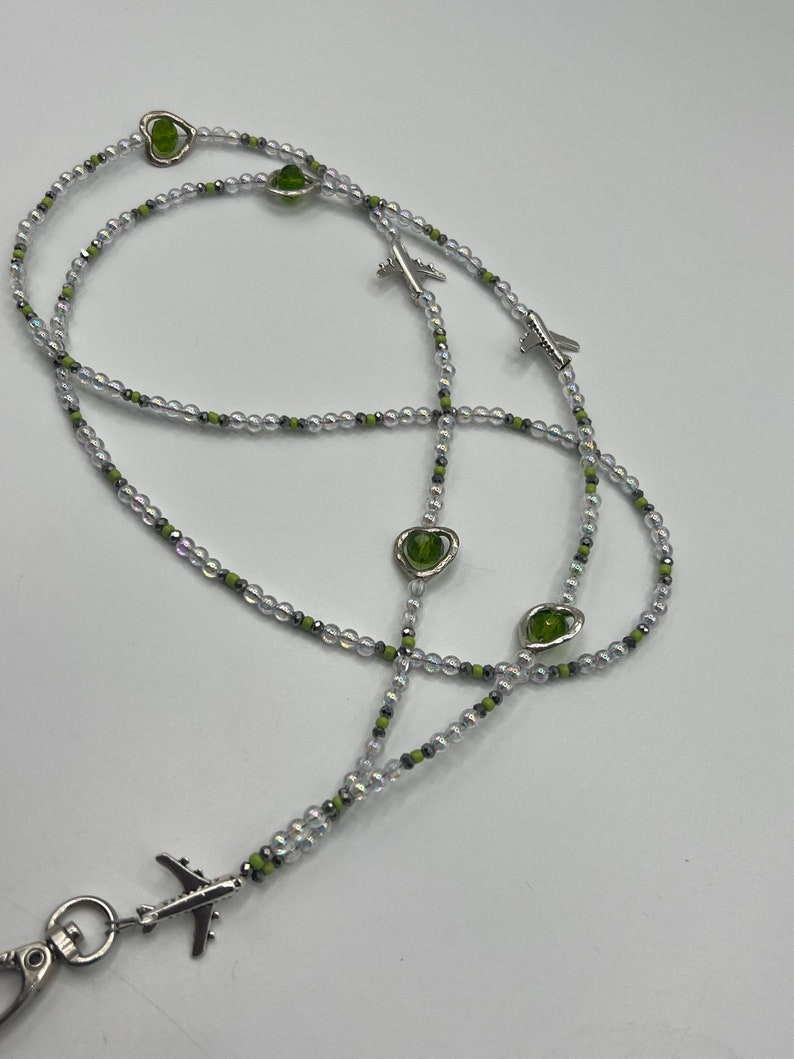 May include: A beaded lanyard with clear, silver, and green beads. The lanyard features silver airplane charms and green heart-shaped beads. A silver clasp is attached at the end.