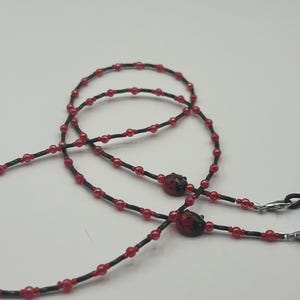 May include: A beaded eyeglass chain with a black cord and red beads. The chain features two ladybug charms and silver-colored clasps for attaching to glasses. The beads are small and evenly spaced along the cord.