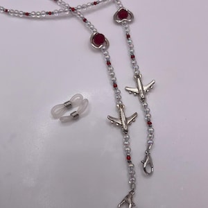 May include: A beaded eyeglass chain with clear and red beads. The chain features silver airplane charms and heart-shaped accents. Includes a pair of clear eyeglass holders with silver metal coils.