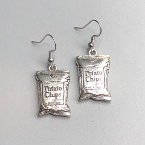 May include: A pair of silver-toned earrings shaped like miniature potato chip bags. Each bag has the words "Potato Chips" printed on the front. The earrings hang from silver-colored fish hook ear wires.