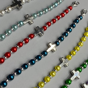 May include: A collection of beaded bracelets in various colors, including white, light blue, red, dark blue, yellow, green, and purple. Each bracelet features a silver-colored cross charm and silver beads. The bracelets are connected with silver clasps.