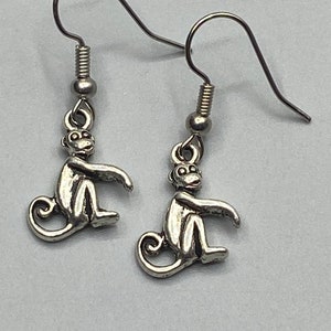 May include: A pair of silver-colored monkey earrings. Each earring features a small, detailed monkey charm with a seated pose, attached to a hook-style ear wire. The monkey charms have a textured appearance, and the earrings are displayed against a light gray background.