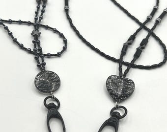 May include: Two black lanyards featuring beaded chains with small star-shaped accents. One lanyard has a circular pendant with a textured silver and black pattern, while the other has a heart-shaped pendant with the same silver and black design.