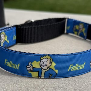 May include: Blue dog collar featuring the word "Fallout" and a cartoon character giving a thumbs-up. The collar has a black nylon strap and a silver metal buckle. The design is repeated around the collar.