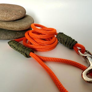 Puede incluir: Una correa naranja para perros con un cierre plateado y asas tejidas de color verde oliva. La correa está enrollada y se encuentra junto a una pila de tres piedras lisas y grises. La correa está hecha de un material tejido duradero.